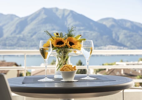 A table with a vase of sunflowers and two glasses of wine, a bowl of snacks, and a view of mountains and water.