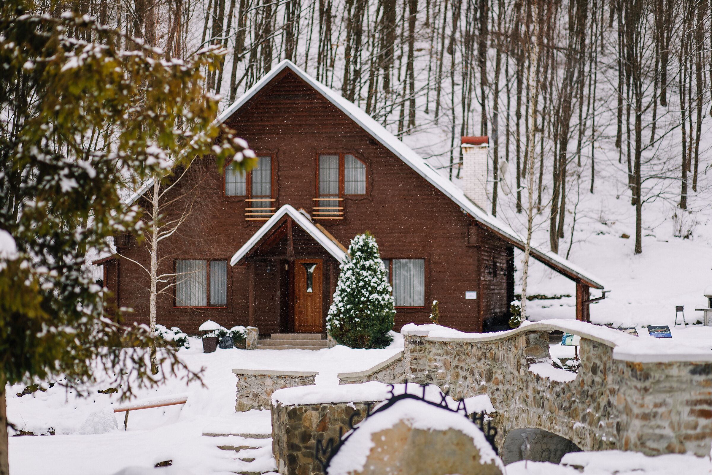 A wooden cabin with a stone entrance, surrounded by snow-covered ground and trees.