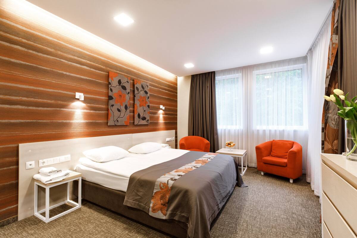 Una habitación de hotel con una cama grande, una mesita pequeña, una silla naranja y un escritorio con una lámpara.