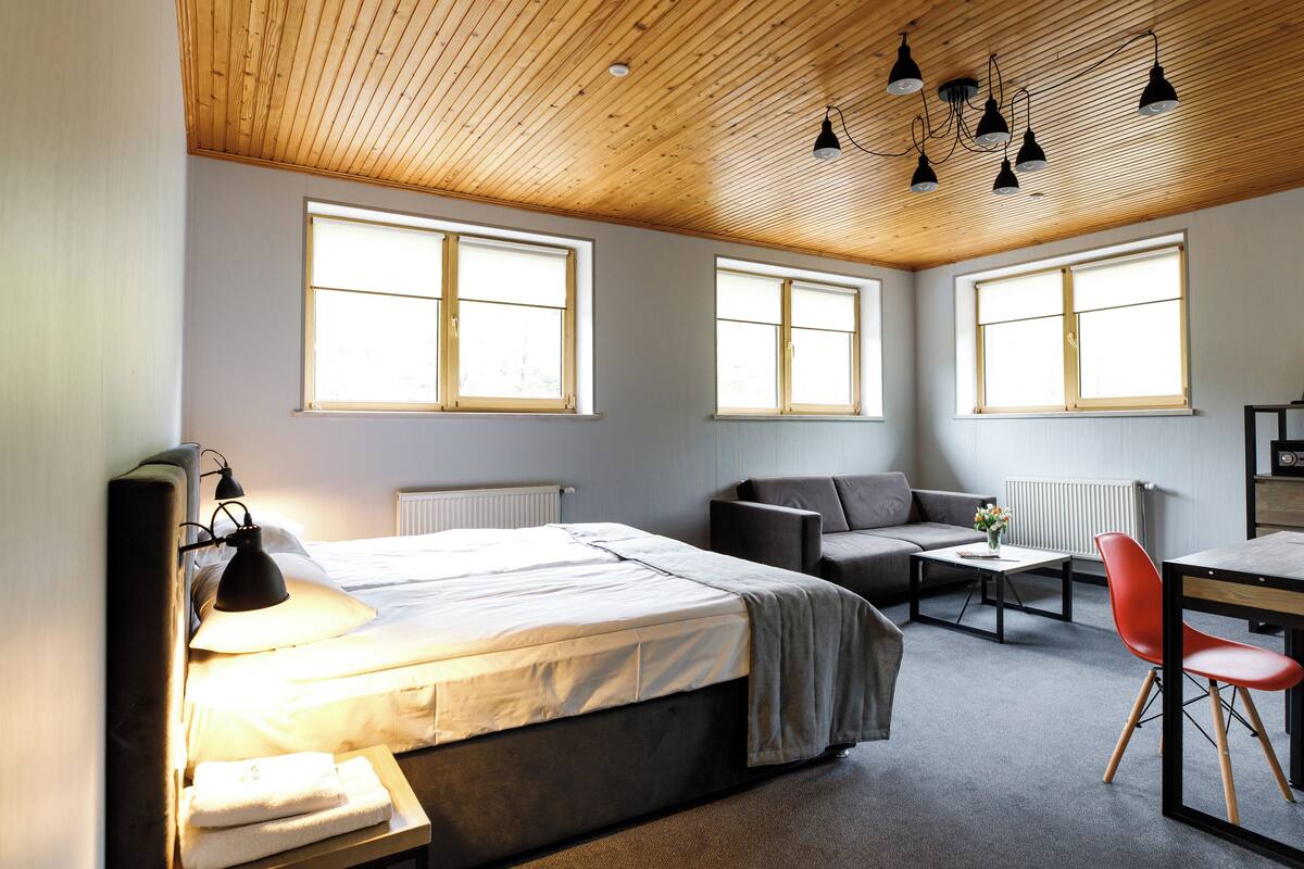 Un dormitorio moderno con una cama, un escritorio y una silla. Amplias ventanas proporcionan luz natural.