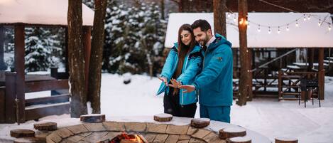 A couple standing by a stone fire pit with snow on the ground and wooden benches in the background.
