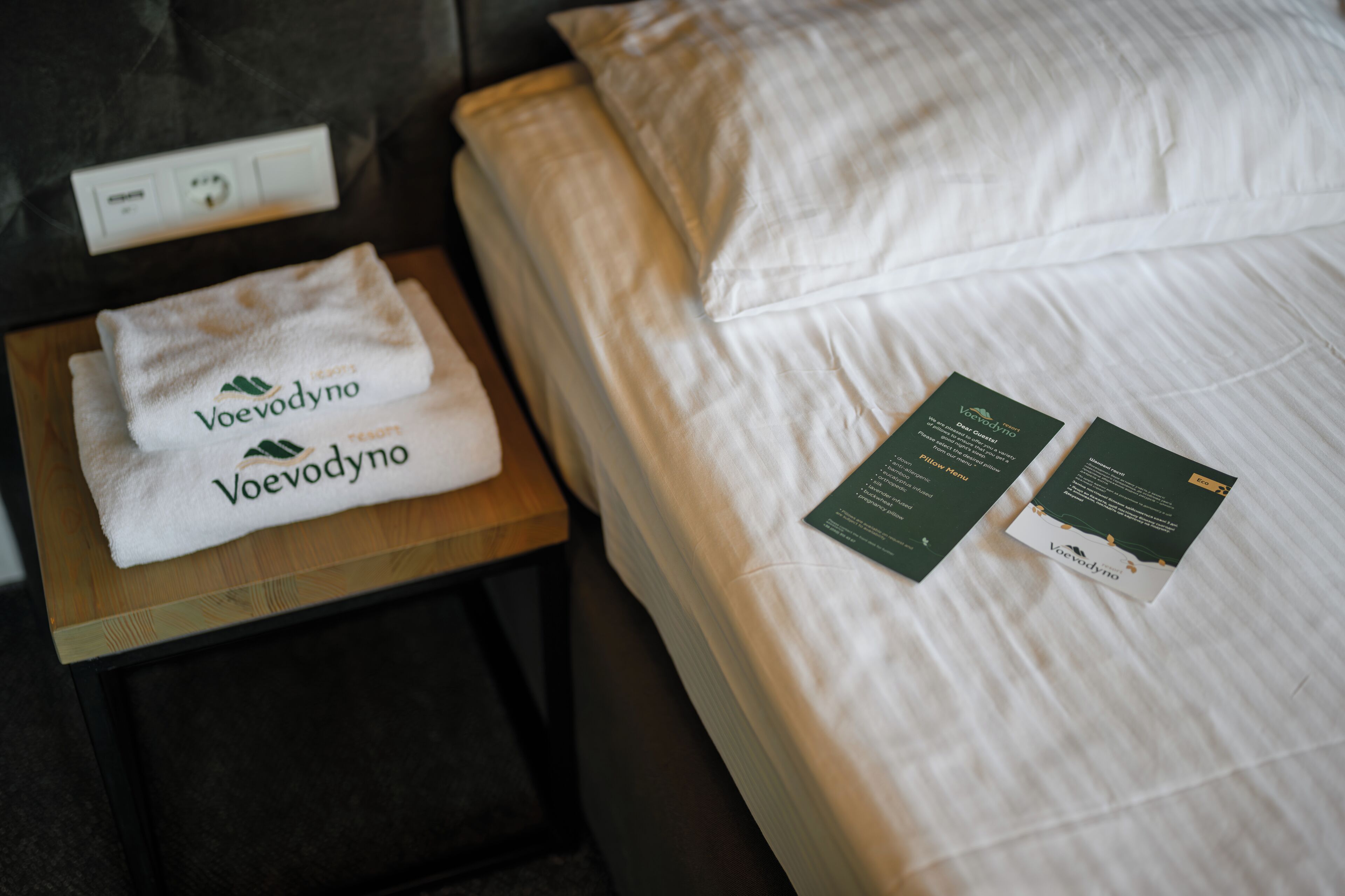 A neatly made bed with white linens and a folded towel on a wooden nightstand. Two informational cards are placed on the bed.