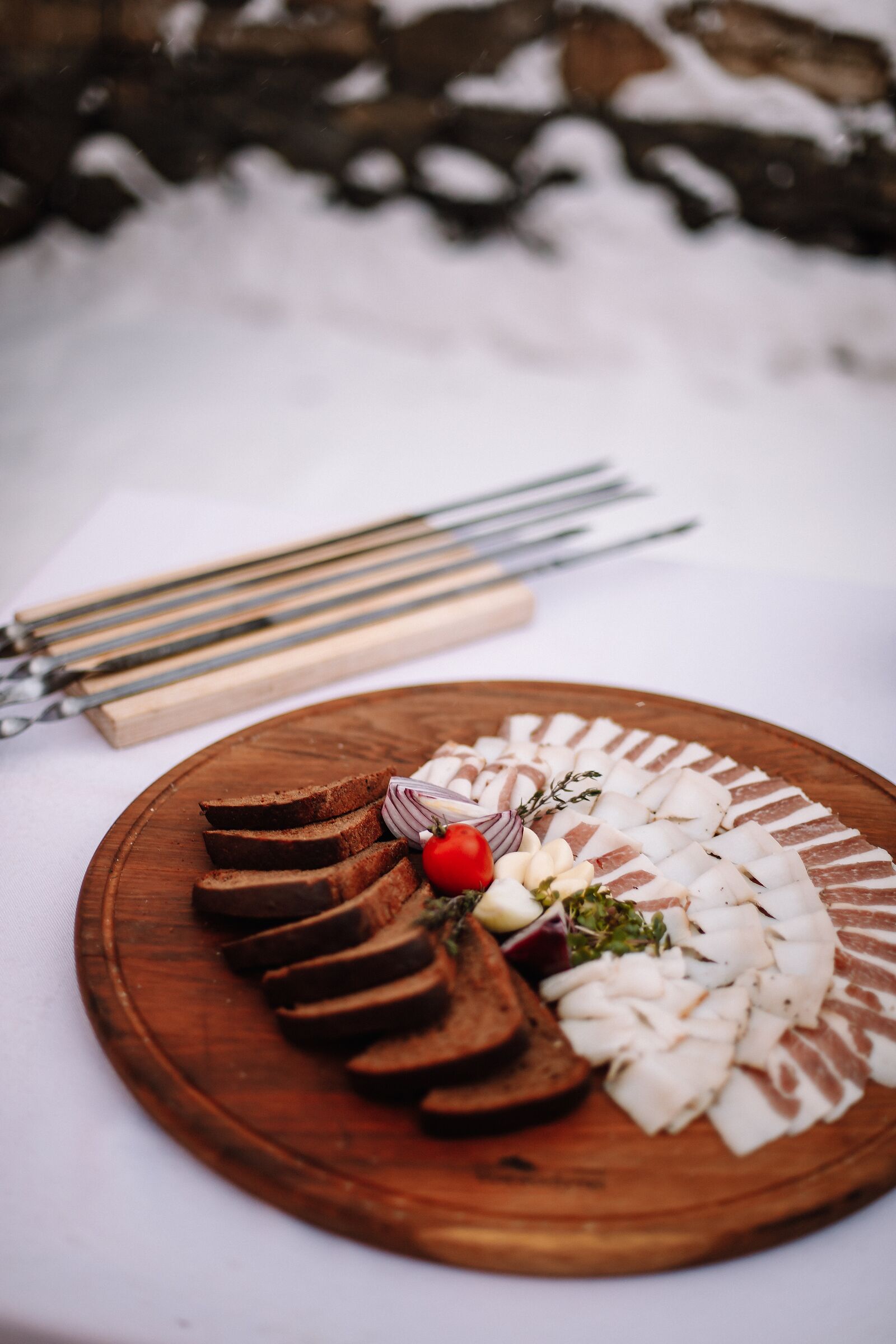 A wooden board with sliced meats, vegetables, and a small red tomato on a white tablecloth.