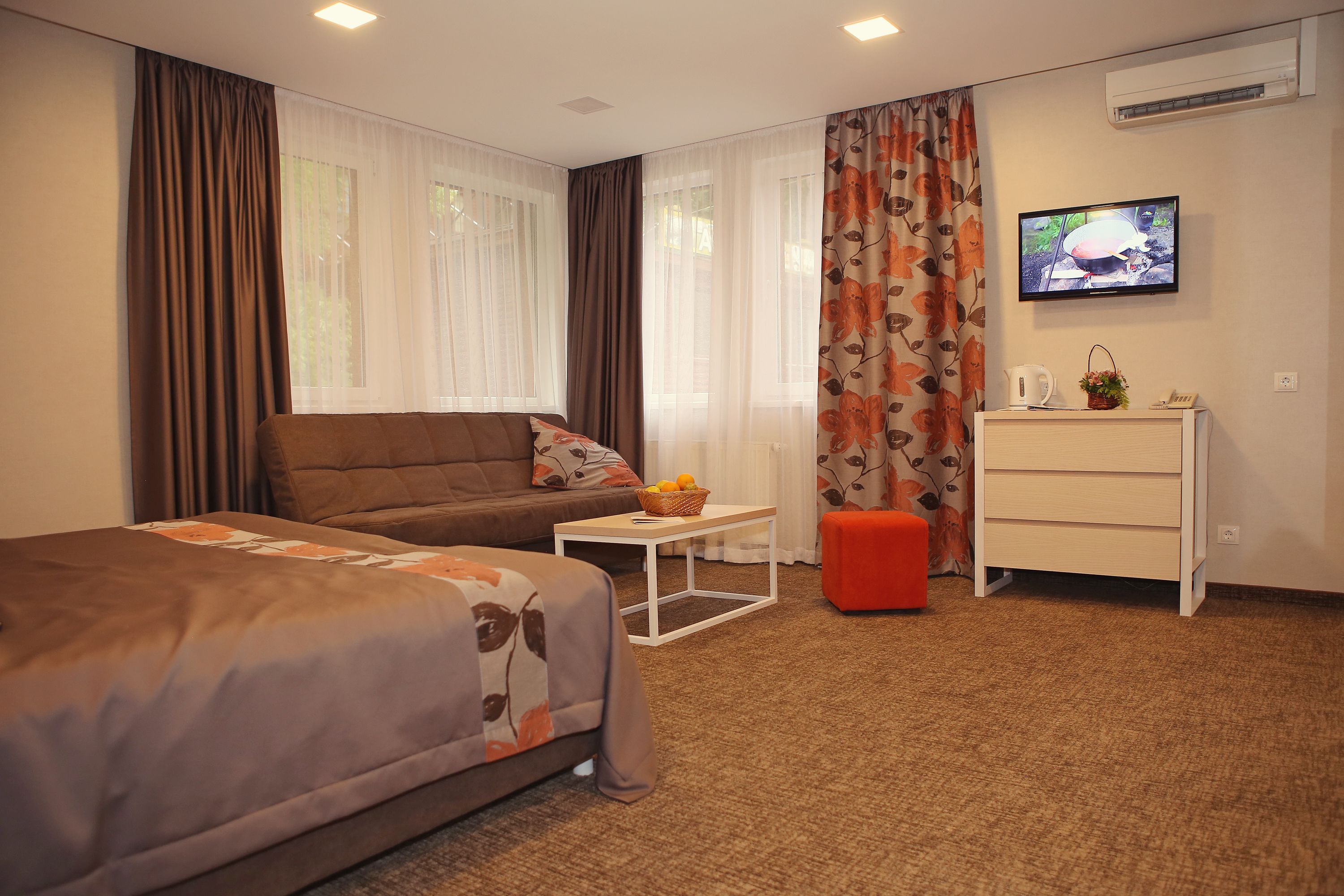 Une chambre d’hôtel avec un lit, un canapé, une petite table sur laquelle se trouve un panier, un pouf rouge, une commode blanche et un téléviseur fixé au mur.