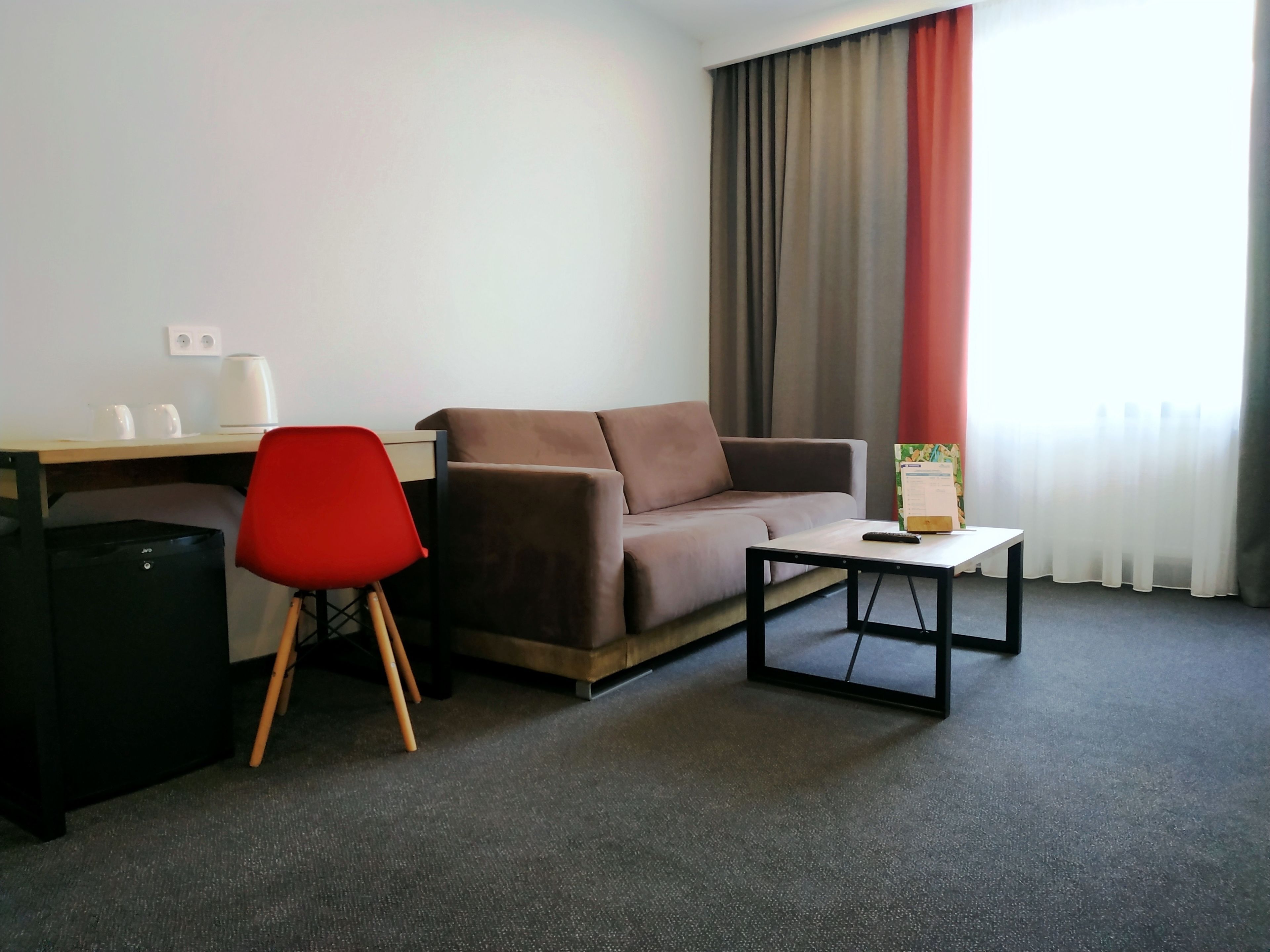 Habitación de hotel con un sofá marrón, una silla roja, una mesita pequeña, un escritorio con tetera y tazas, y un cubo de basura negro.