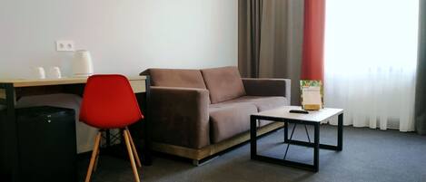 Habitación de hotel con un sofá marrón, una silla roja, una mesita pequeña, un escritorio con tetera y tazas, y un cubo de basura negro.