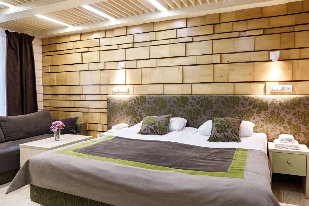 Una habitación de hotel con cabecera de madera, cama con ropa de cama verde y blanca, mesita de noche con lámpara y sofá con un jarrón de flores.