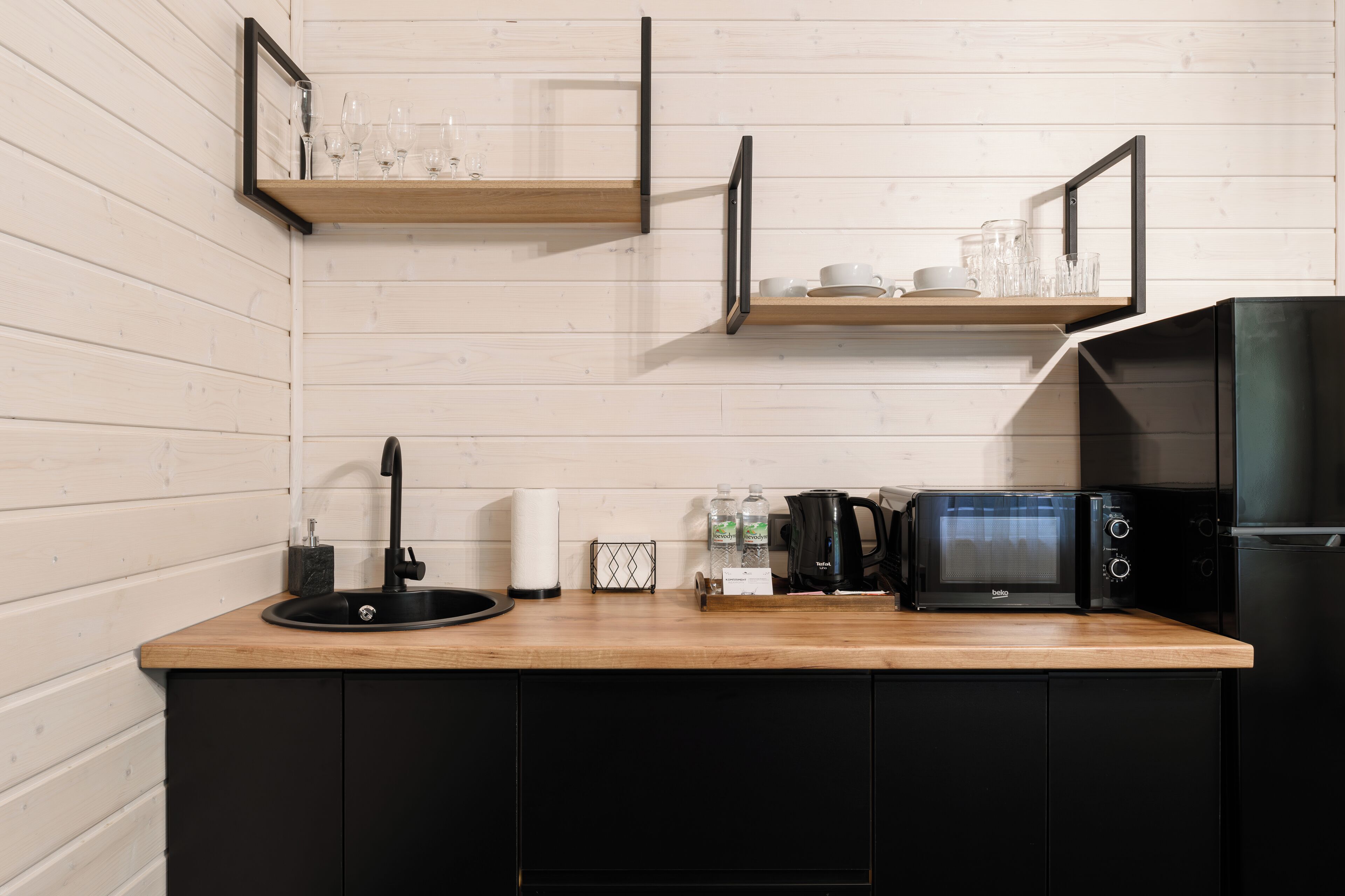 Una cocina con armarios de madera, encimera negra, un fregadero, un microondas y dos estantes con cristalería.