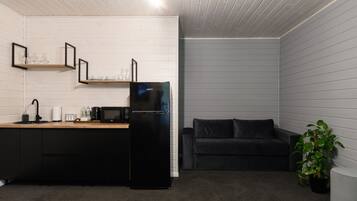 Cocina compacta con un refrigerador negro, un microondas y una cafetera sobre una encimera de madera. En la sala de estar, hay un sofá negro y una planta en maceta.