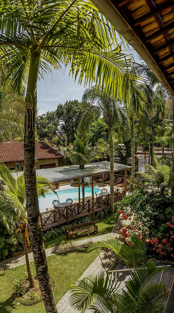 A pool area with a wooden deck, palm trees, and a view of a traditional building with a tiled roof. - Pousada Casarão da Praia (São Sebastião)