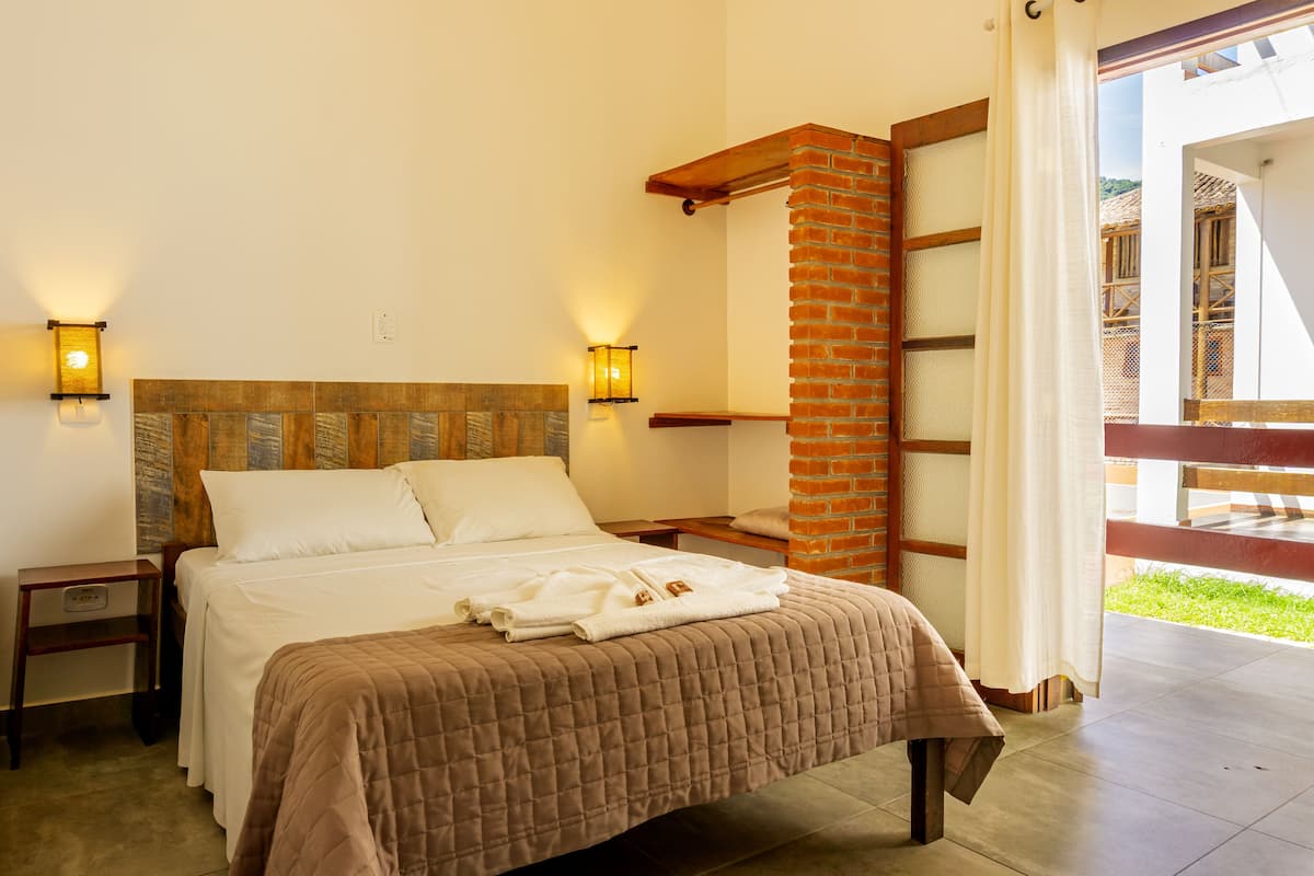 A bedroom with a bed, two bedside tables, a brick accent wall, and a view of the outside.
