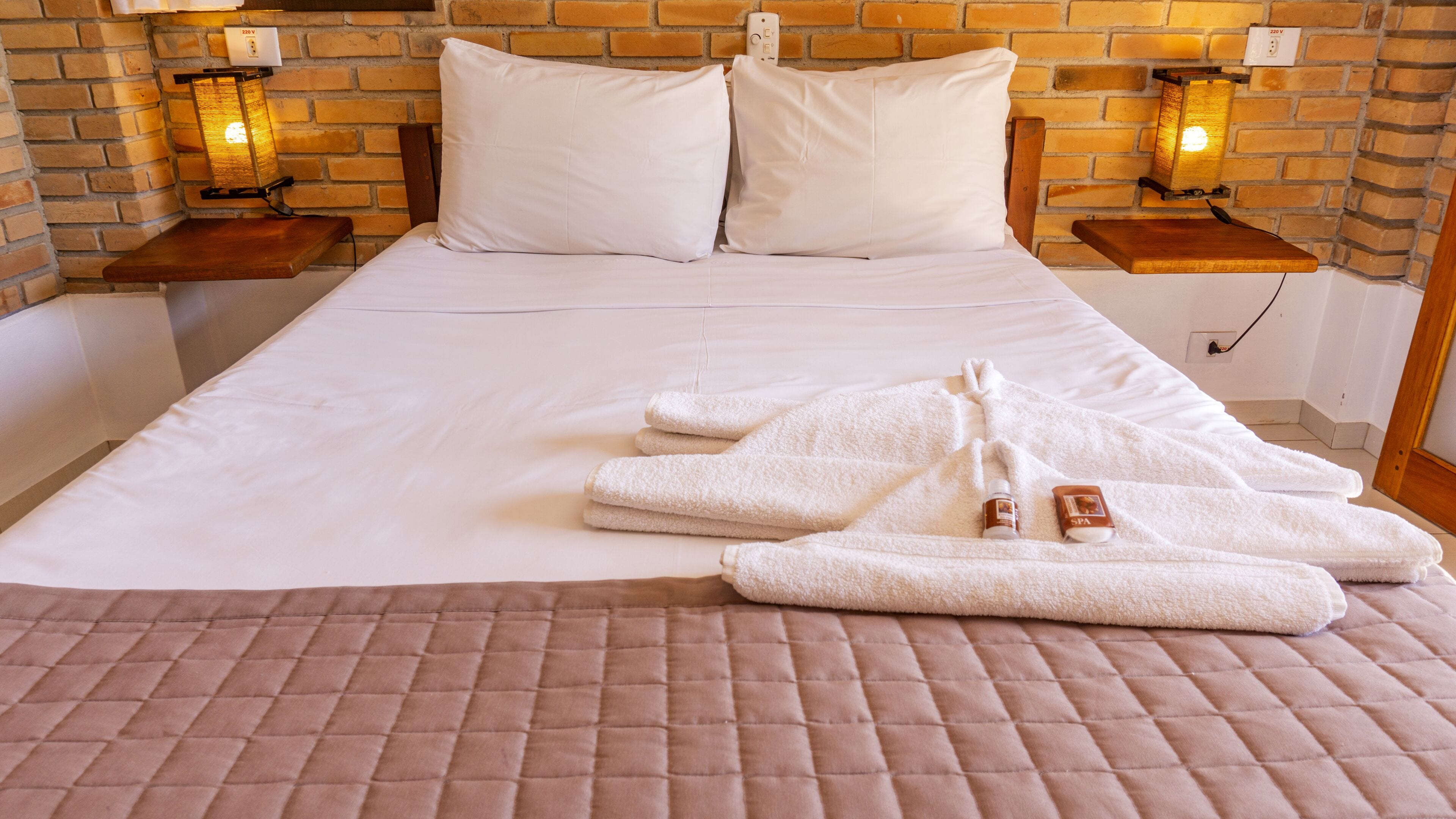 A neatly made bed with white linens, two pillows, and a quilted brown bedspread. There are two bedside tables with lamps, and a small bottle and container on the bed.