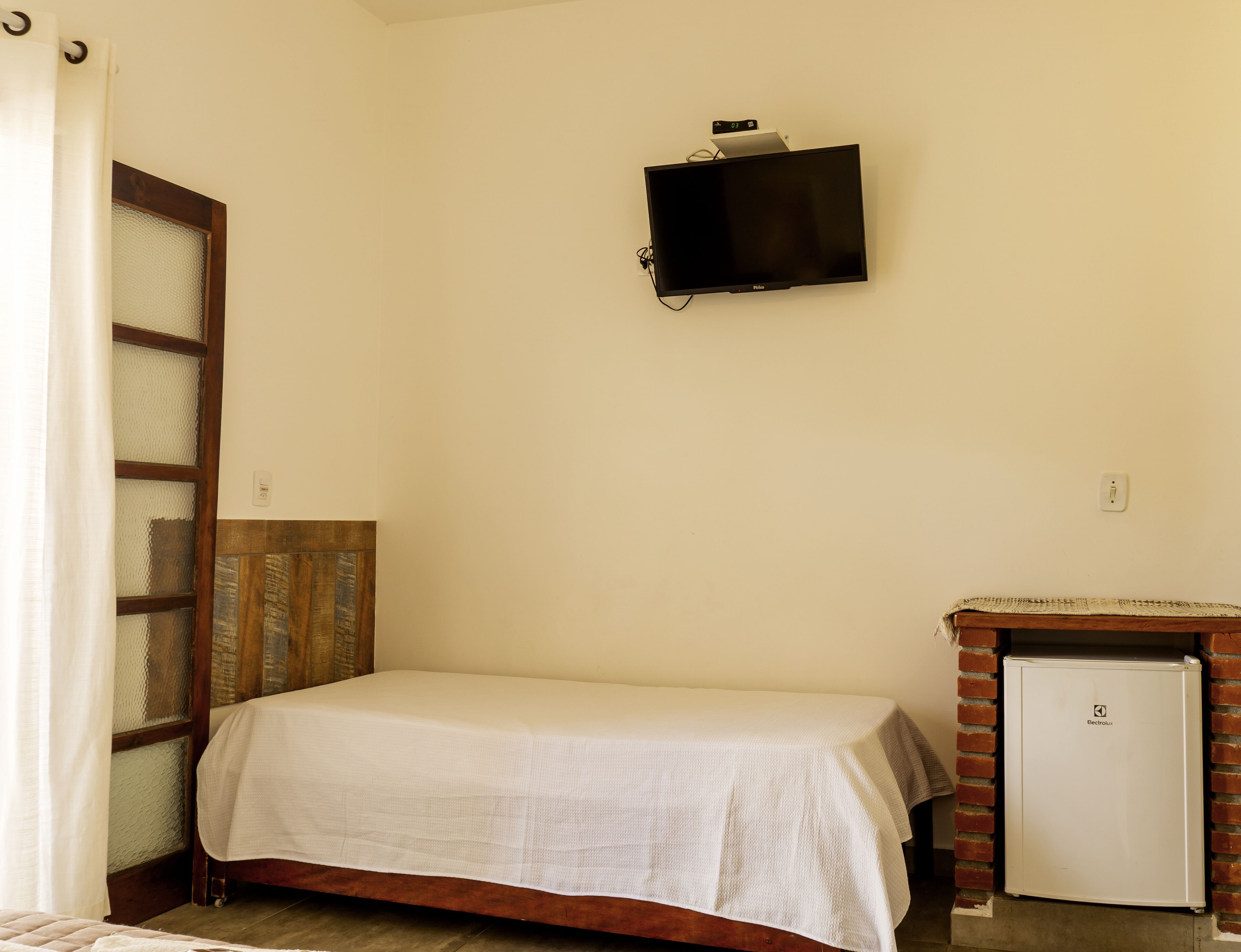 A small, simple room with a bed, a television mounted on the wall, and a refrigerator.