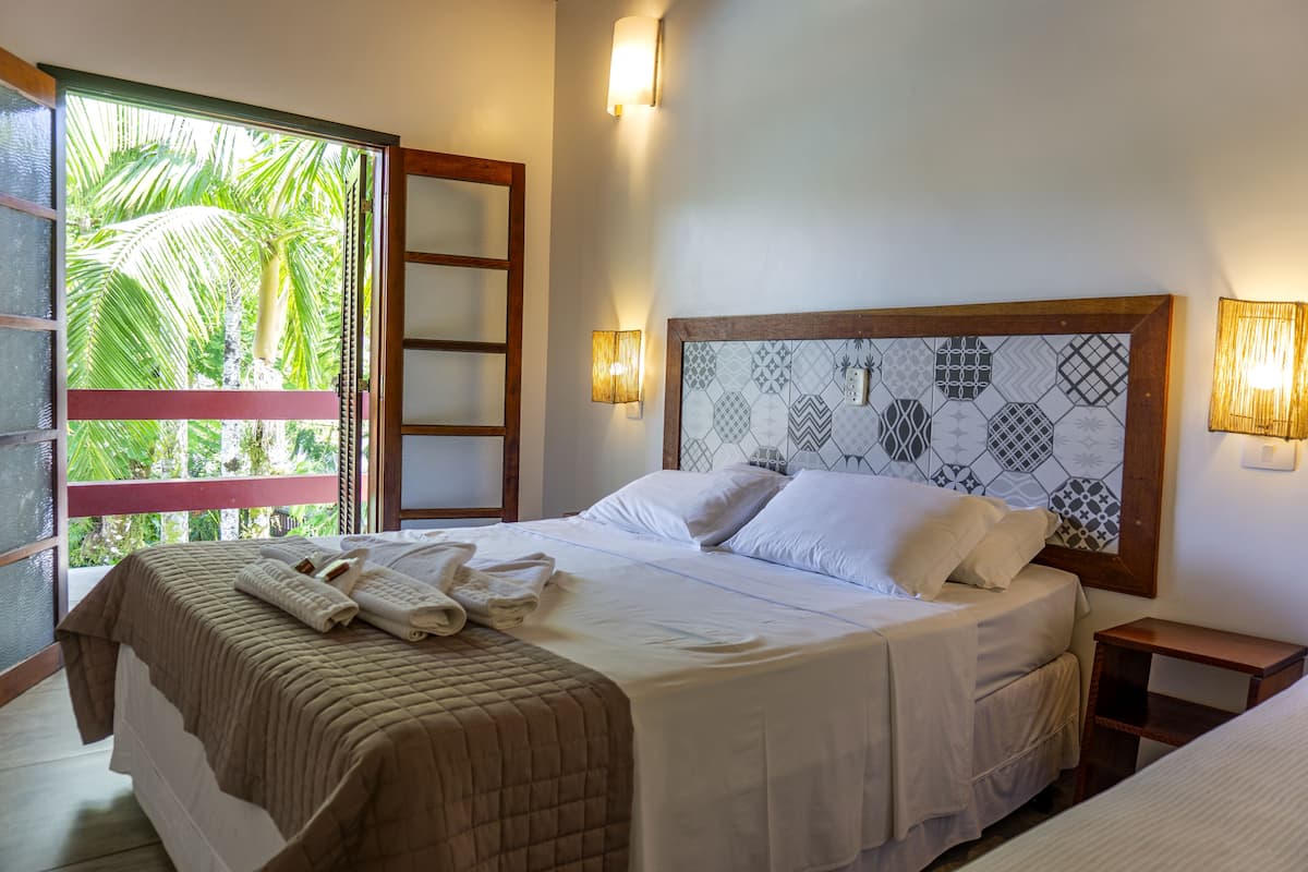 A neatly made bed with a patterned headboard, a bedside table, and a view of palm trees through a glass door.