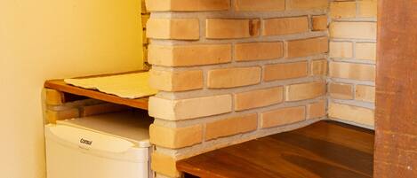 A corner of a room with a brick wall, a wooden shelf, and a small white refrigerator.