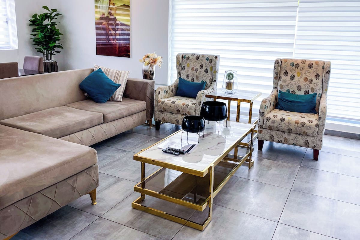 Un salon moderne avec un canapé beige, deux fauteuils à motifs, une table basse en verre et une petite table d’appoint.
