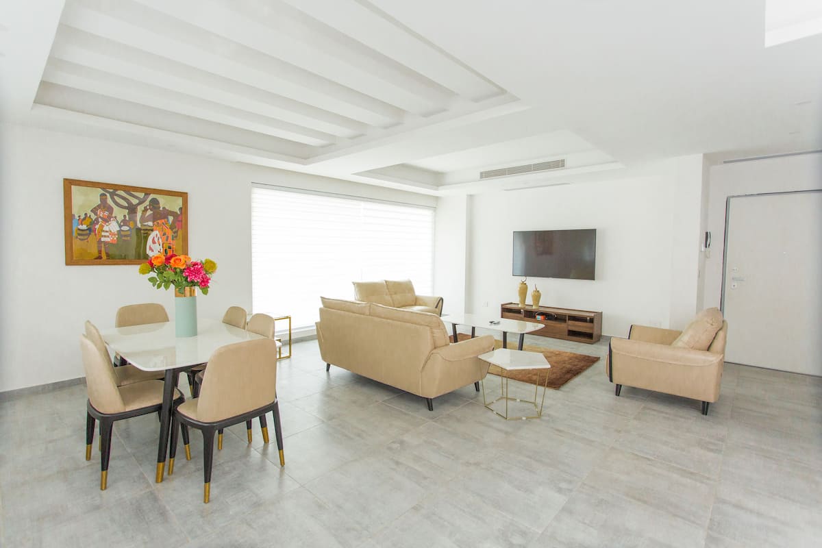 Un salon moderne comprenant un coin repas, des canapés de couleur beige, une table basse et un téléviseur à écran plat fixé au mur.