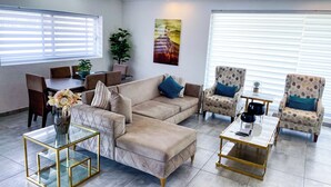 Een moderne woonkamer met een beige bank, twee fauteuils, een glazen salontafel en een eethoek met een tafel en stoelen.
