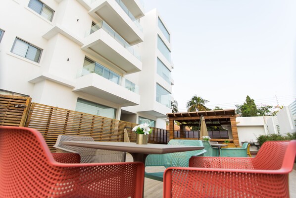 Un espace extérieur moderne aménagé avec des chaises rouges et une table, une clôture en bois et, au fond, un bâtiment doté de balcons.