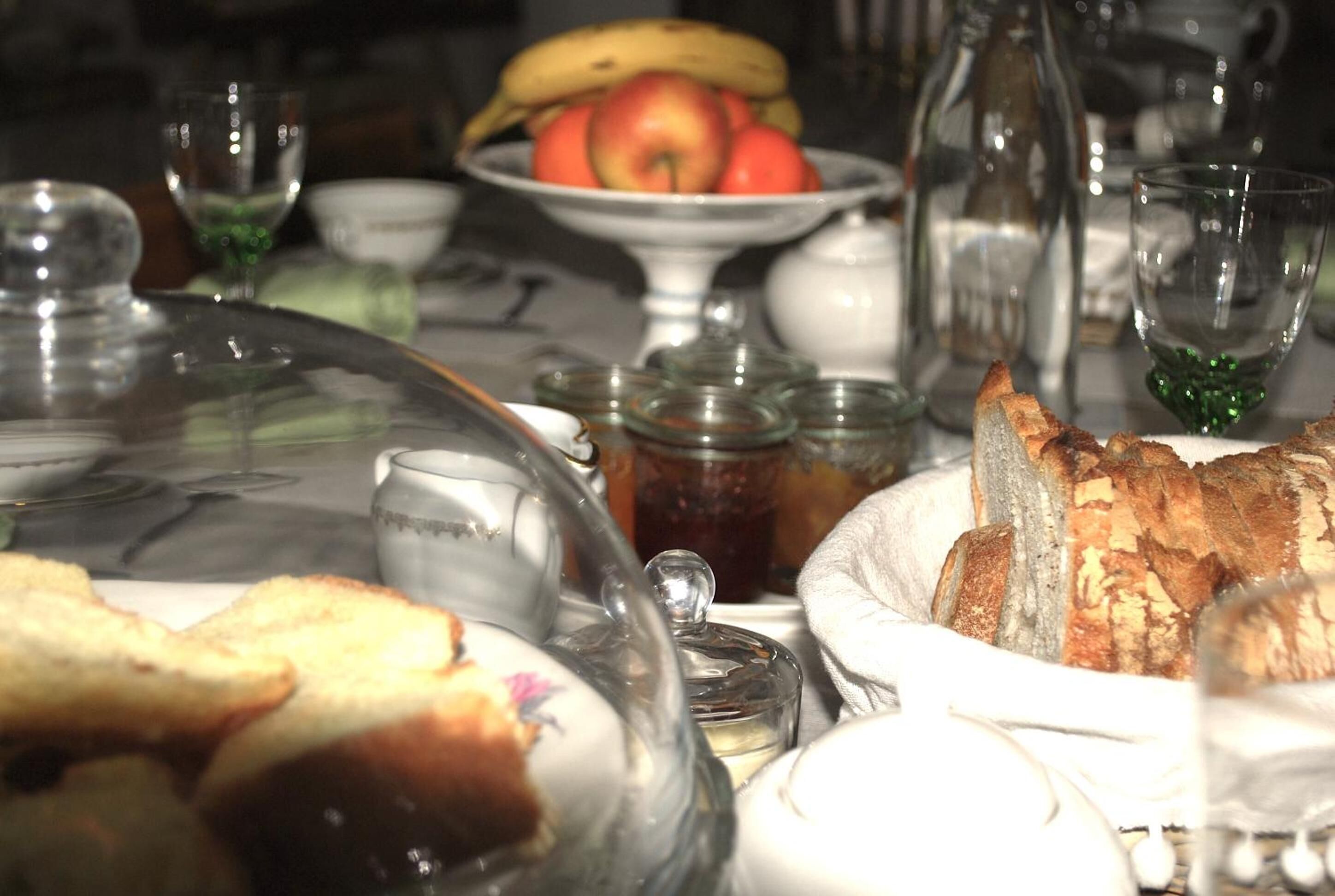 A dining table with a variety of food items, including bread, a bowl of fruit, and a glass container.