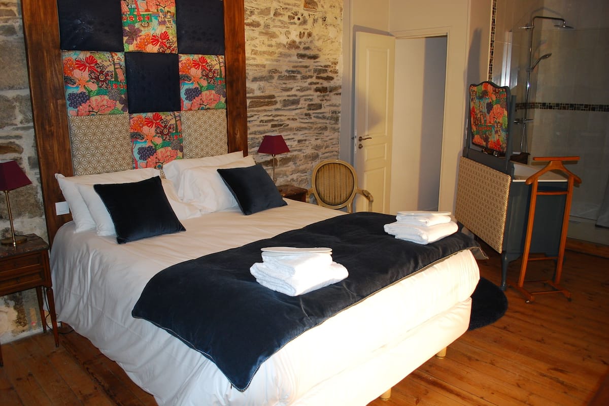 A neatly made bed with white and dark blue linens, a wooden headboard, and a stone wall behind it.
