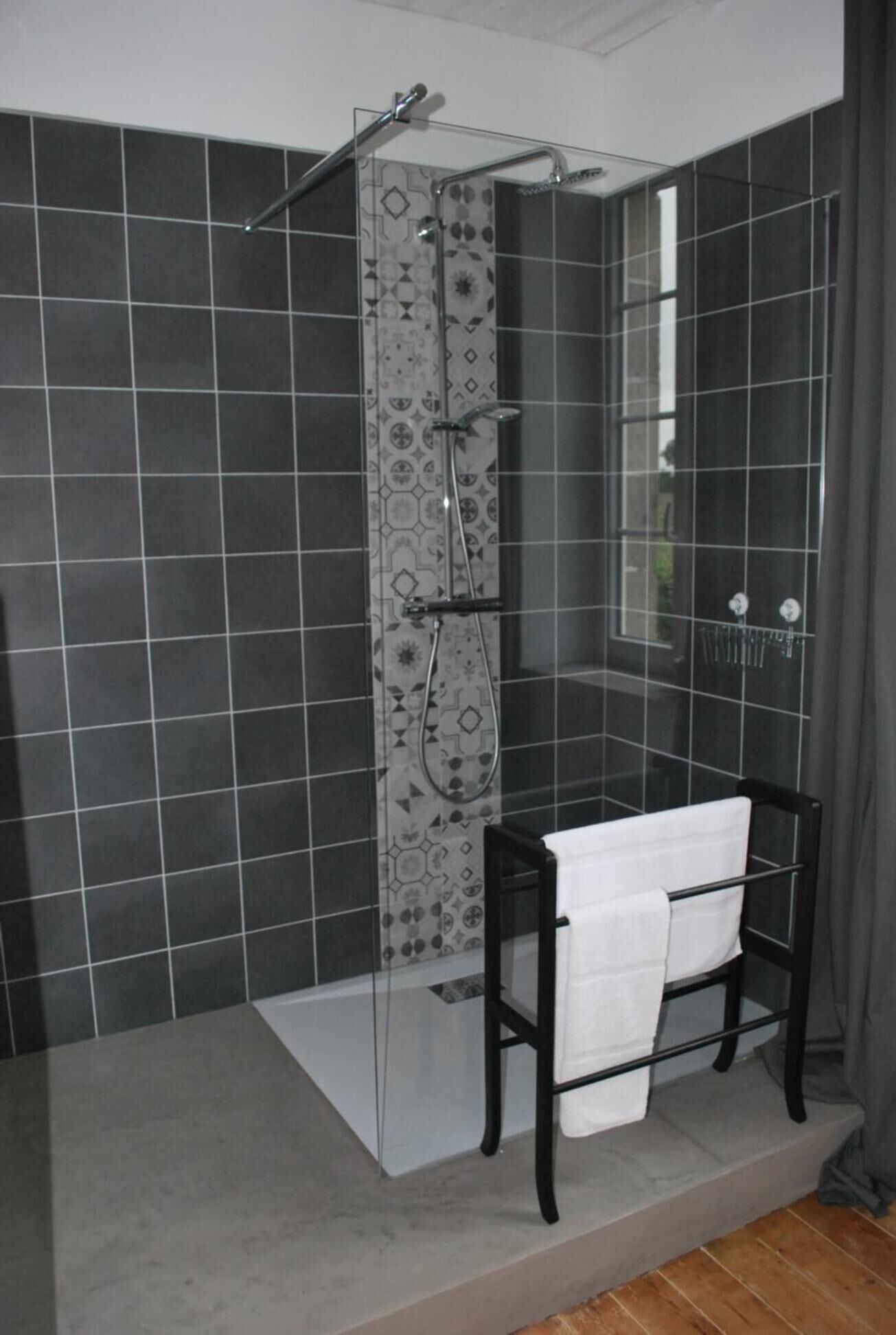 A bathroom with dark tiled walls, a glass shower enclosure, a black sink with a white towel, and a window with curtains.