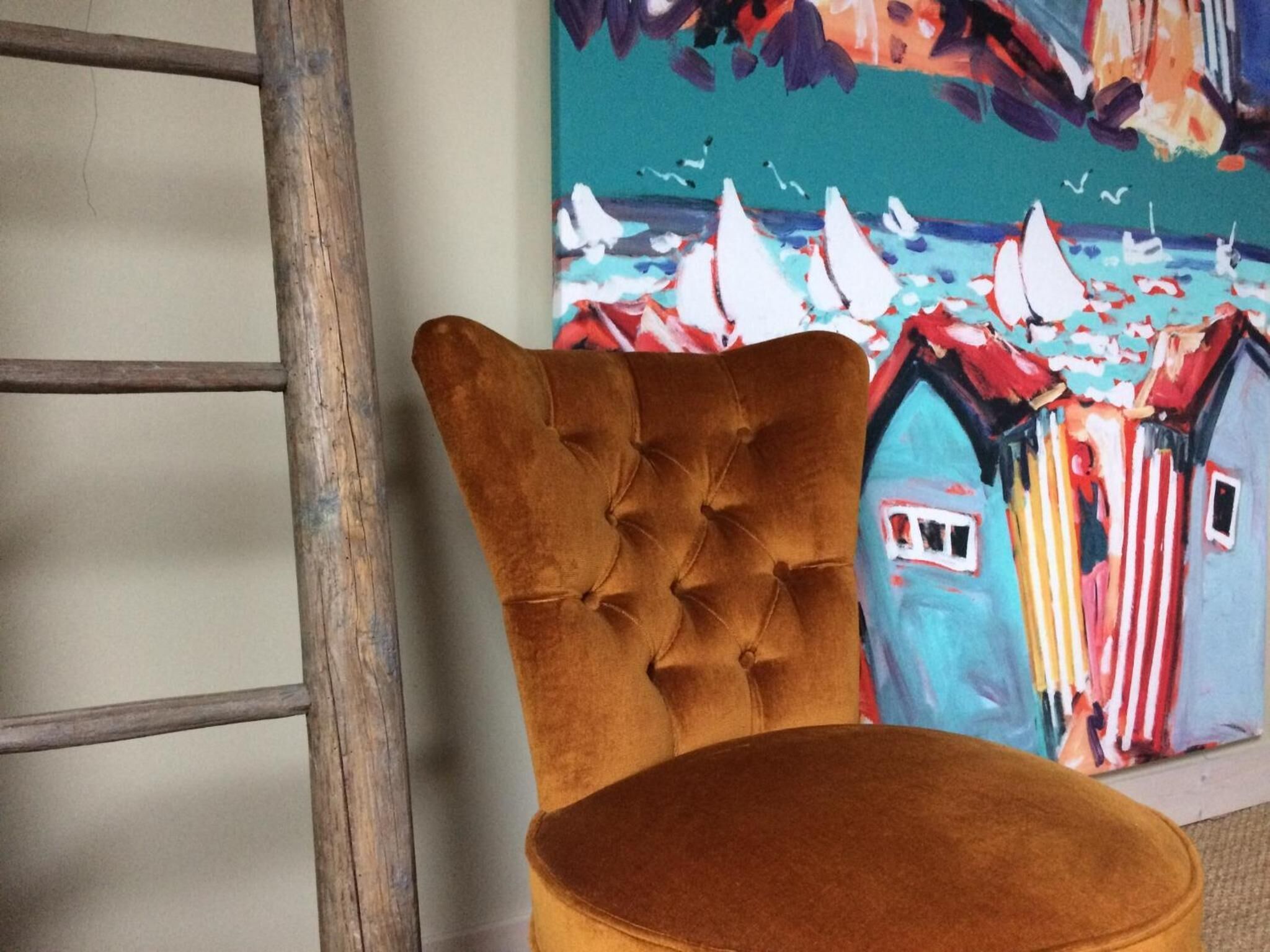A chair with a tufted backrest and a cushion, positioned next to a colorful painting of a coastal scene with sailboats.