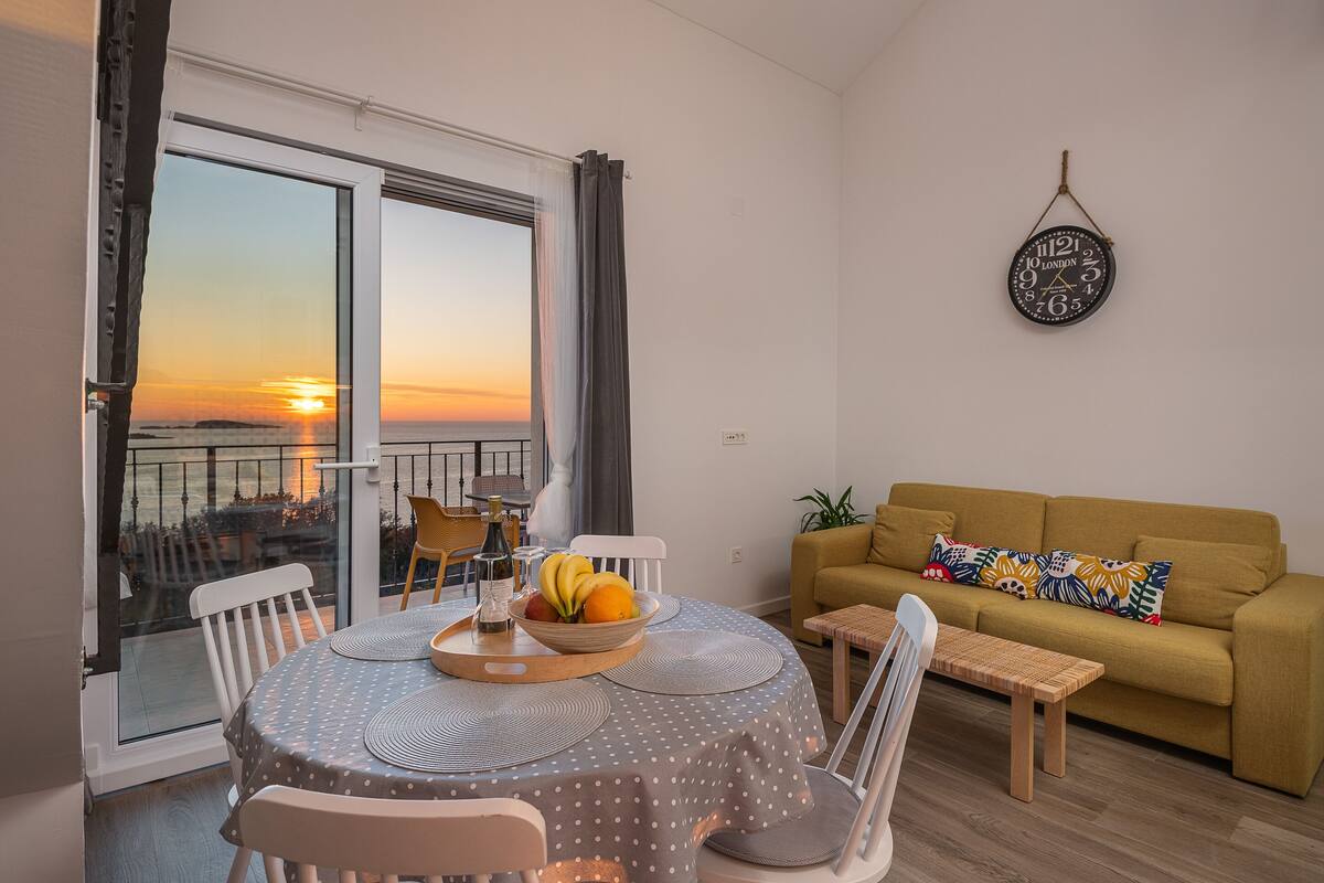 Ein Essbereich mit Tisch für zwei Personen, einer Couch und Blick auf den Sonnenuntergang durch eine Balkontür.