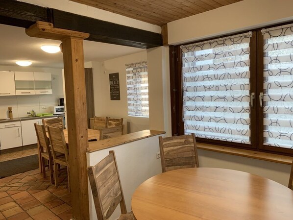 Un coin repas avec une table et des chaises en bois, une cuisine à l’arrière-plan et des stores à motifs aux fenêtres.