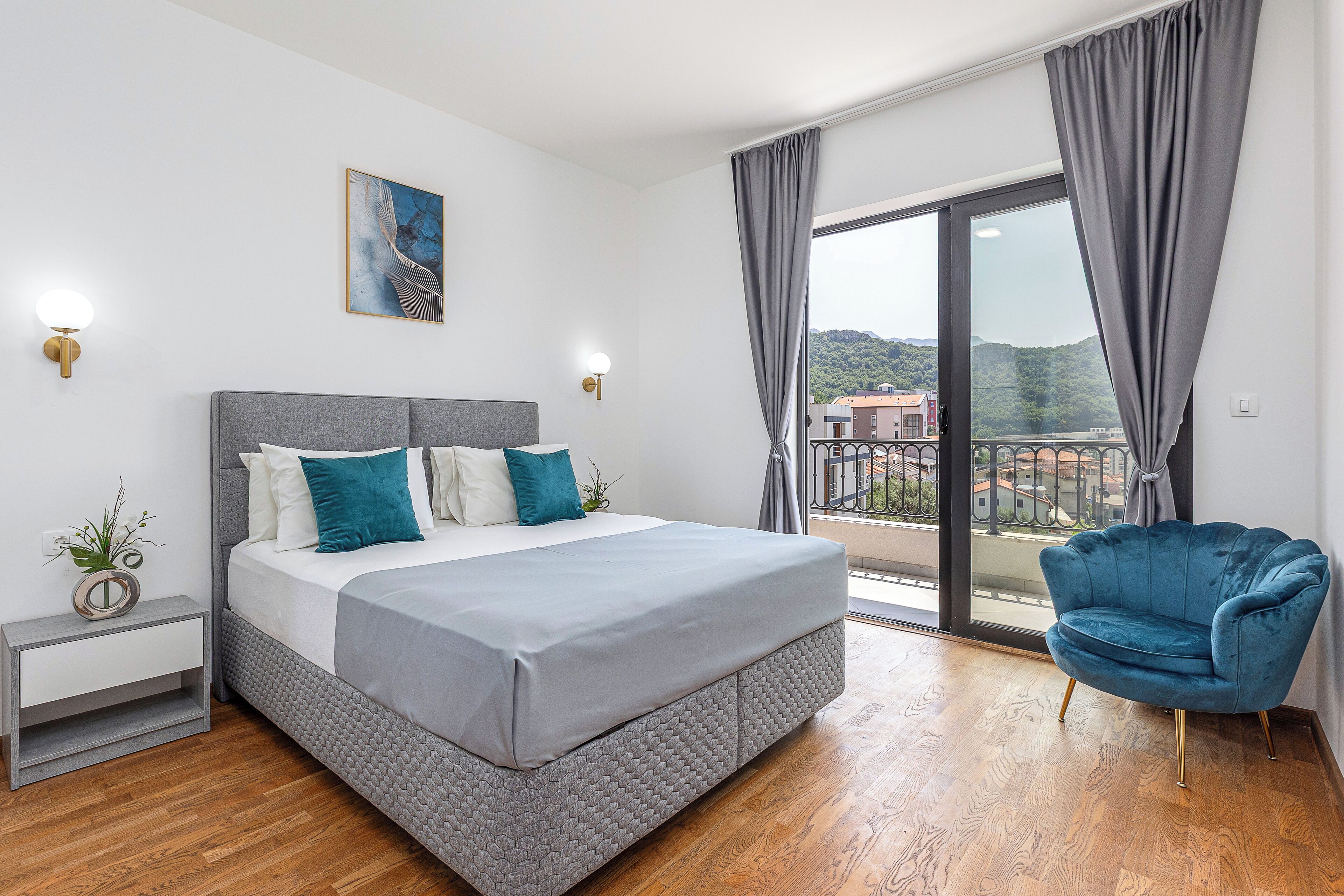 Un dormitorio moderno con una cama grande, una mesita de noche, un sillón azul y un balcón con vistas a la ciudad.