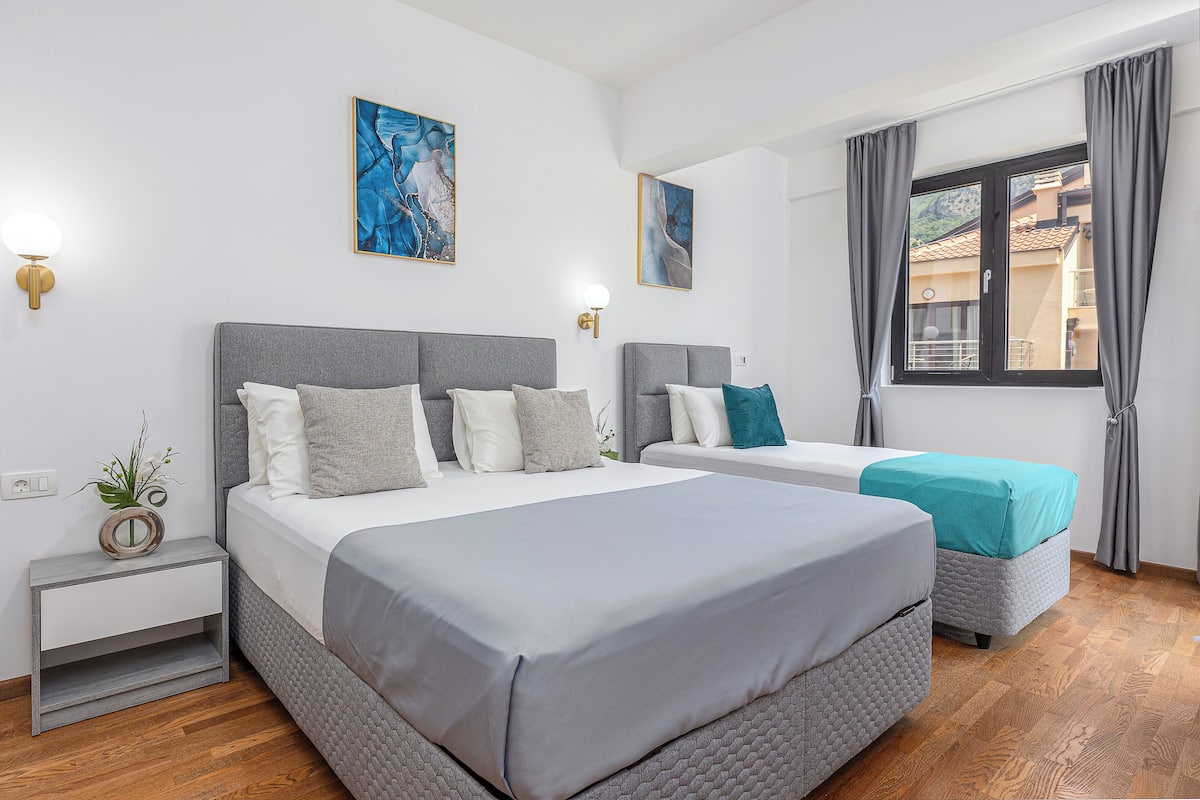 A hotel room with two beds, a nightstand, a wall-mounted lamp, and a window with curtains.