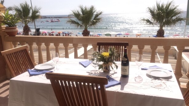 A table set for two with a bottle of wine, a vase with flowers, and a view of the beach with umbrellas and people in the distance. - Beautiful apartment facing the sea with huge terrace (El Campello)