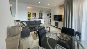 Ein modernes Wohnzimmer mit einer Couch, einem Couchtisch und einem Fernseher. Im Hintergrund befindet sich eine Küchenzeile.