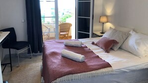 Ein Hotelzimmer mit einem Bett, einem Schreibtisch, einem Stuhl, einer Lampe und Blick ins Freie durch eine Glastür.