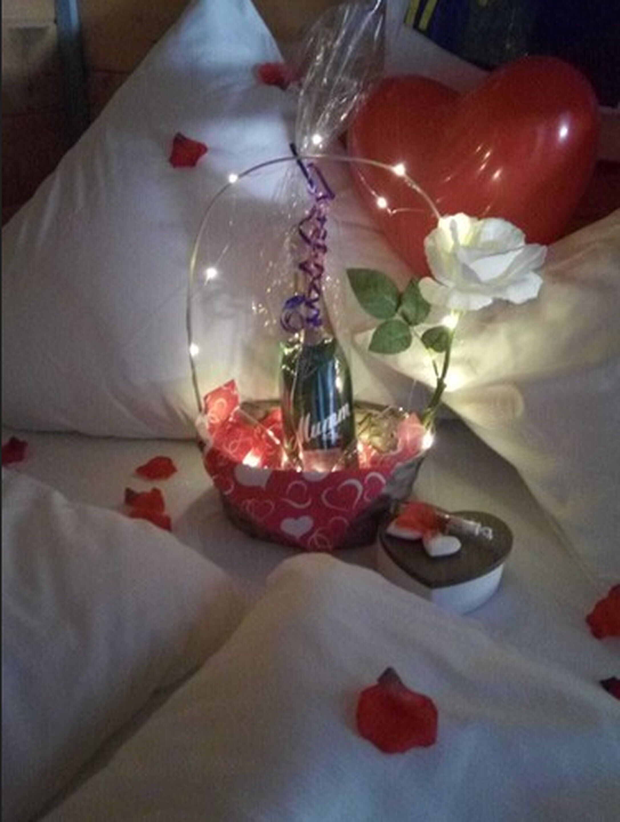 Decoração com uma garrafa transparente, uma rosa branca e um balão em formato de coração sobre uma cama com almofadas brancas e pétalas de rosa vermelhas.