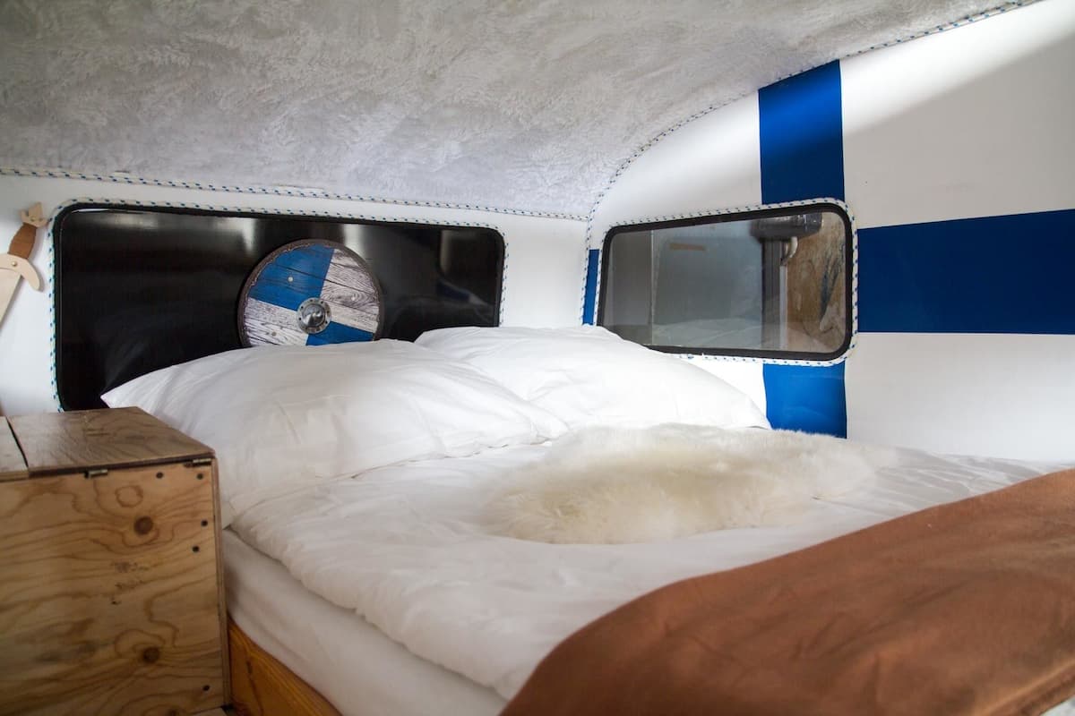 A compact bed with a white mattress and a brown blanket, a wooden box, and a fan with a blue and white design.