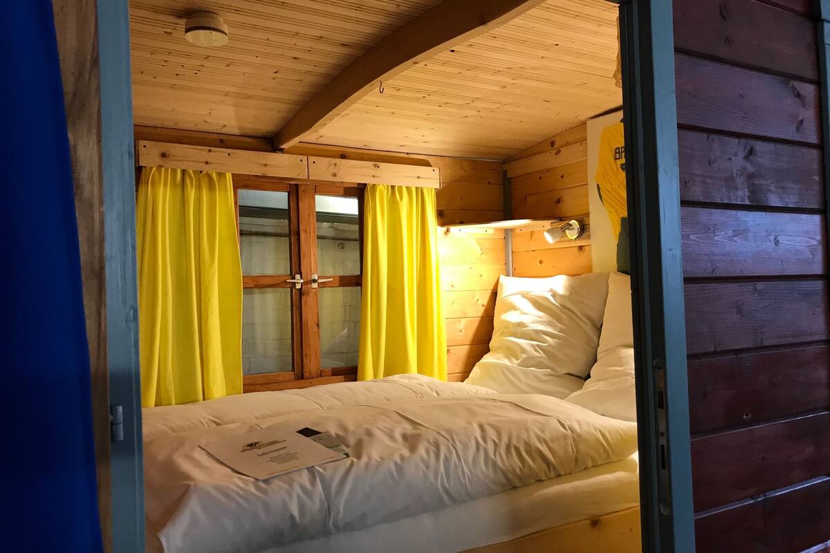 A wooden cabin room with a bed, yellow curtains, and a window with a wooden frame.