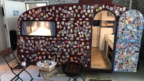 A room with a camper-style setup, featuring a large window and a wall covered in various stickers. There is a small table with a basket and a cup, and two chairs. - HafenTraum IndoorHostelCamp (Bremen)
