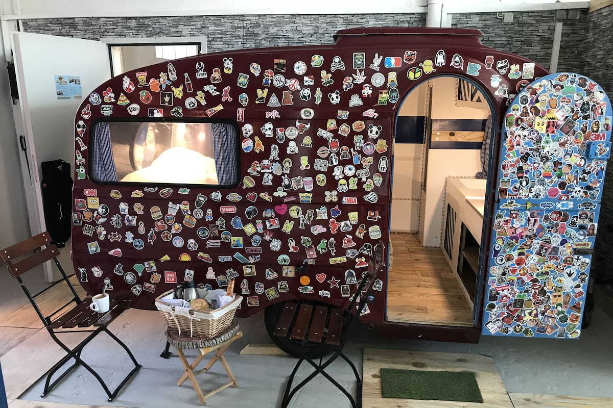 A room with a camper-style setup, featuring a large window and a wall covered in various stickers. There is a small table with a basket and a cup, and two chairs.