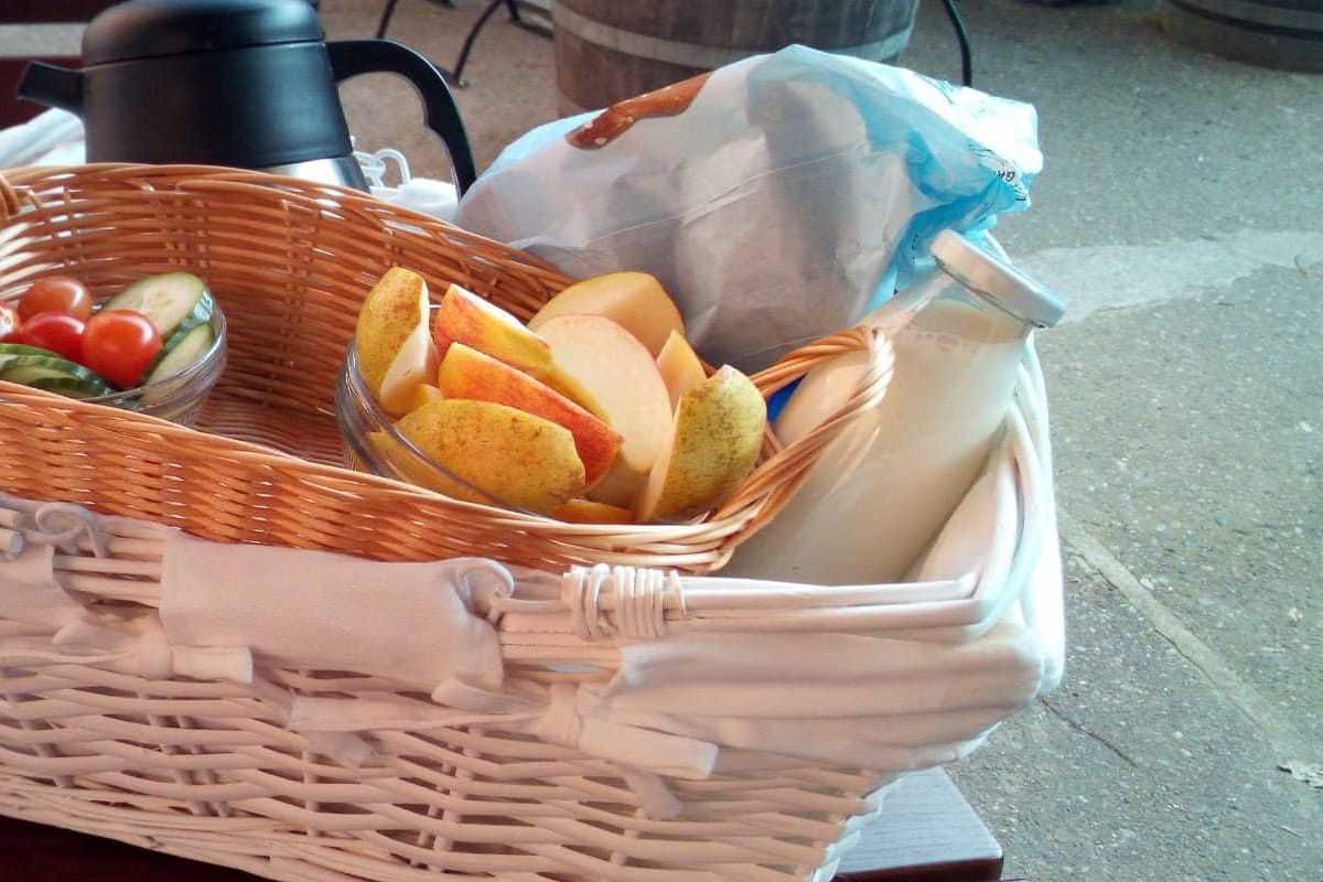 A wicker basket containing fruits, vegetables, and a bag of bread on a wooden bench.