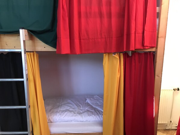 Una cama litera con una base inferior y una escalera, una cama individual con ropa de cama blanca y cortinas en rojo, amarillo y verde.