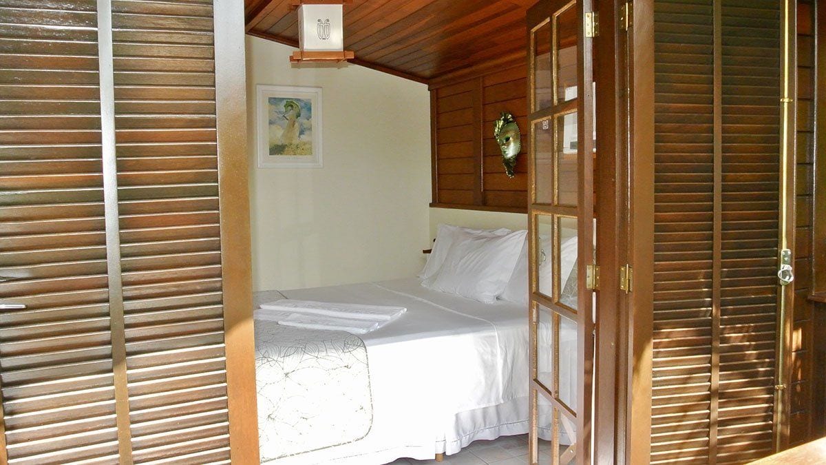A room with a bed, wooden shutters, a painting on the wall, and a small light fixture.