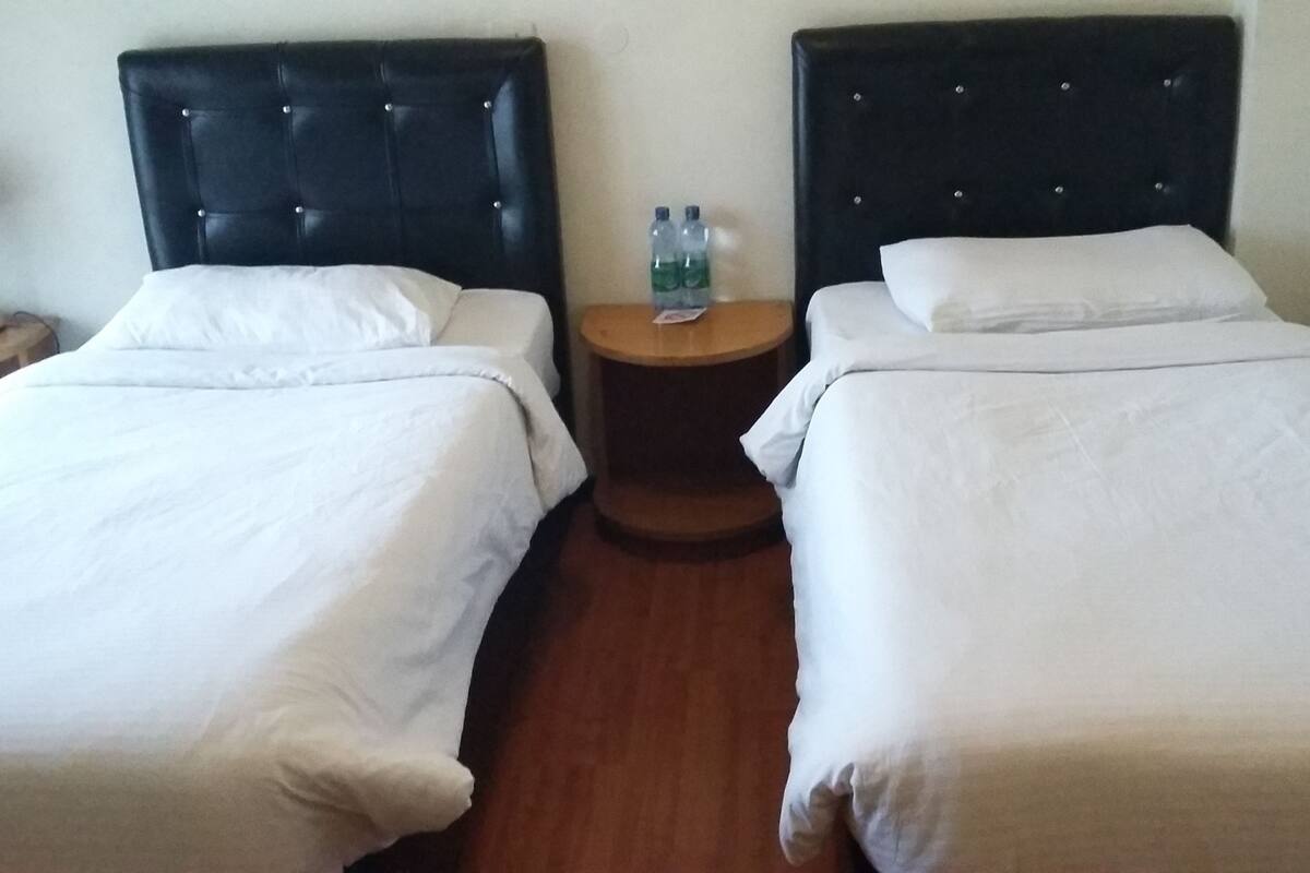A hotel room with two single beds, a round wooden nightstand, and two bottles of water on the nightstand.