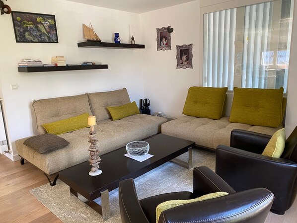 A modern living room with a sofa, coffee table, and chairs. There are shelves with decorative items and a window with blinds.