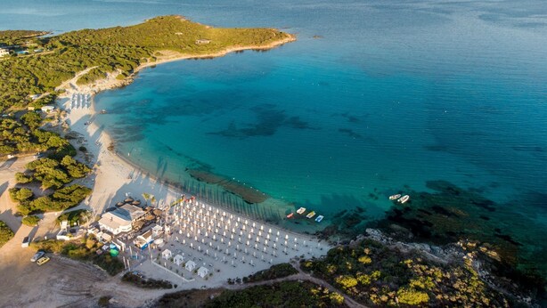 Luchtbeeld van een kustgebied met een strand, een resort met rijen parasols, en een bergachtige achtergrond.