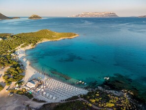 Vista aérea de una zona costera con playa, un complejo turístico con hileras de sombrillas y un paisaje montañoso al fondo.