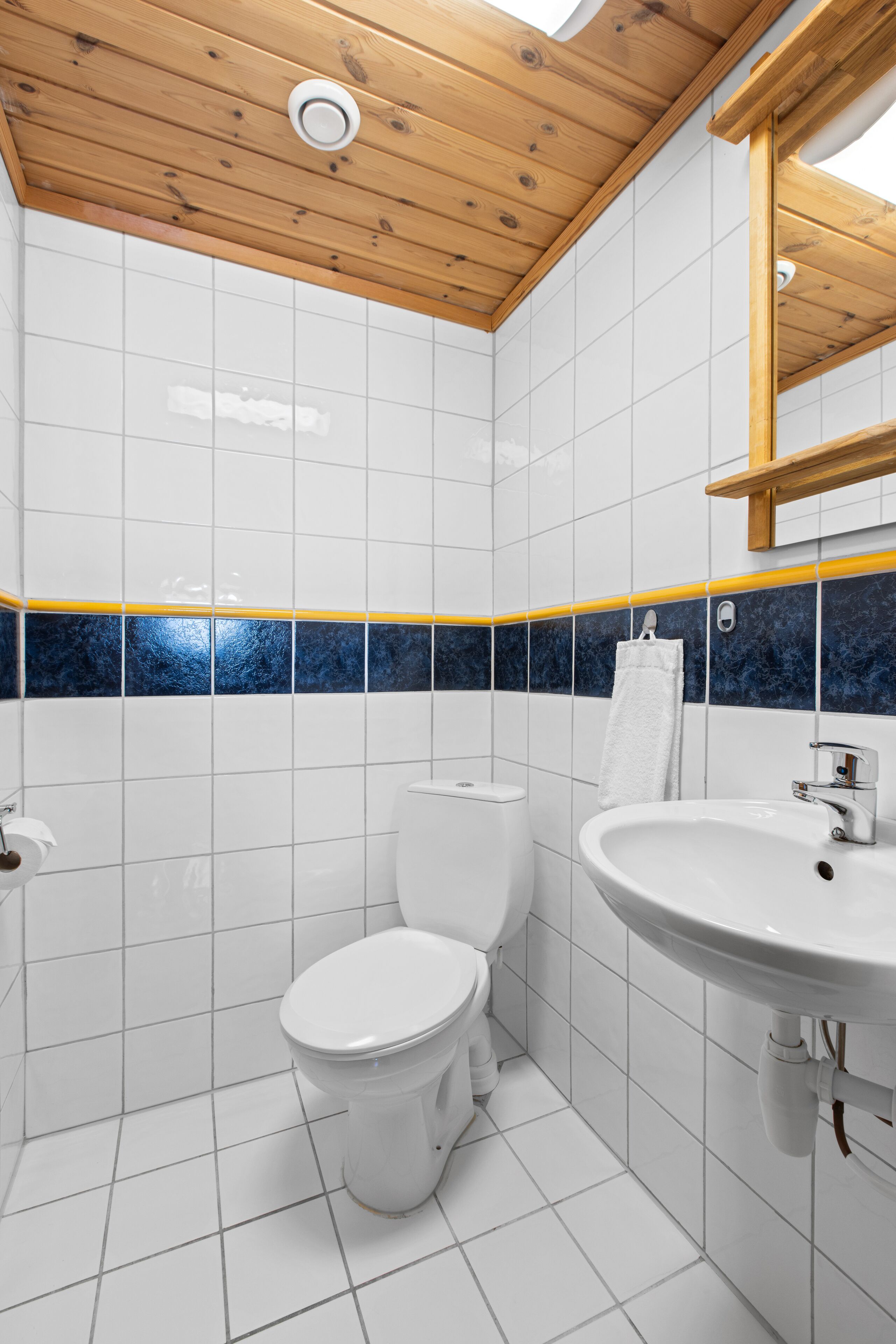 Een badkamer met witte tegels, een houten plafond, een accentmuur in blauw en geel, een wit toilet en een wastafel met spiegel erboven.