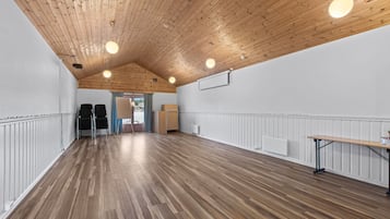 A spacious room with a wooden ceiling and white walls, featuring a long table and chairs along one wall.