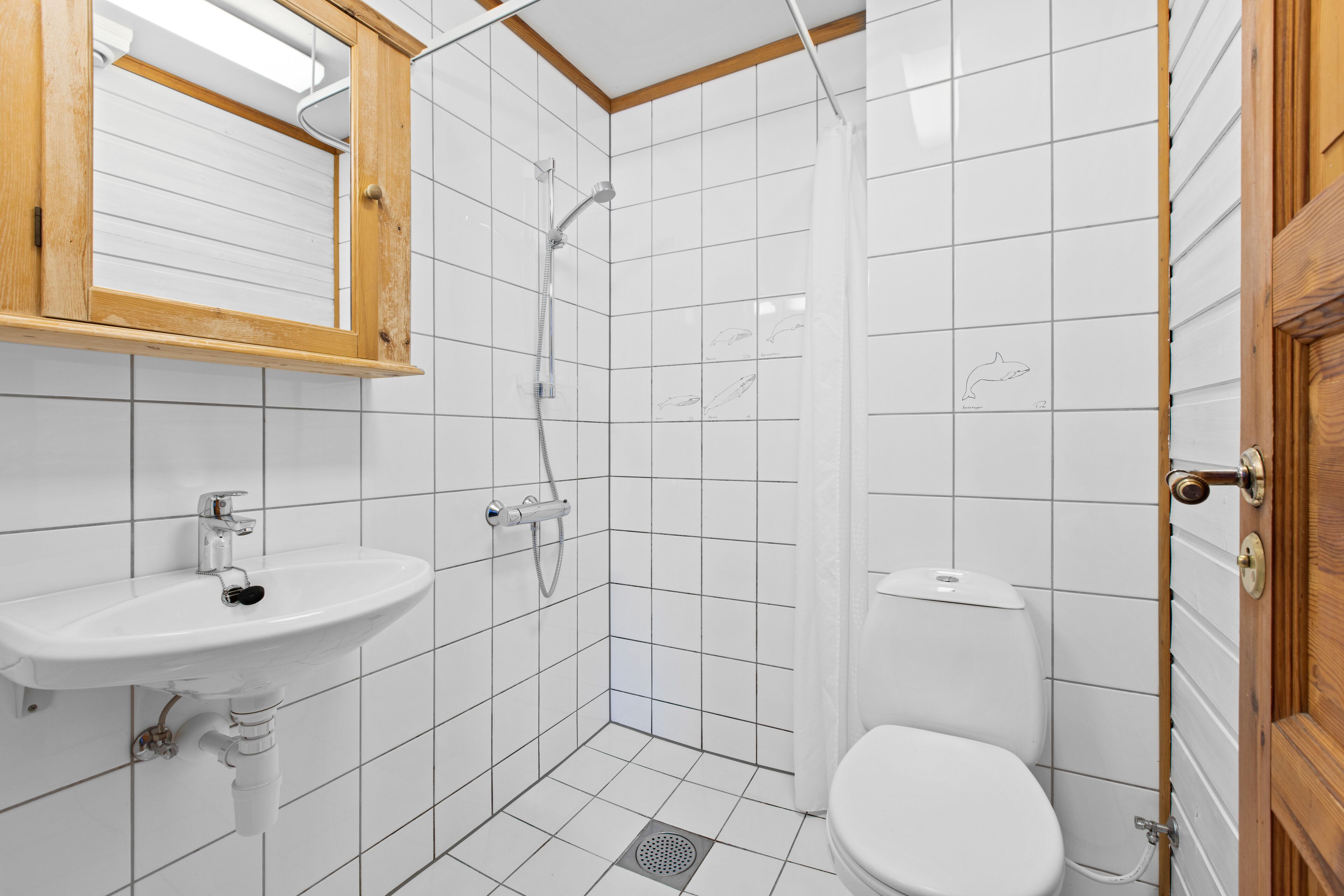 A bathroom with a white sink, shower, and toilet, wooden door, and tiled walls.