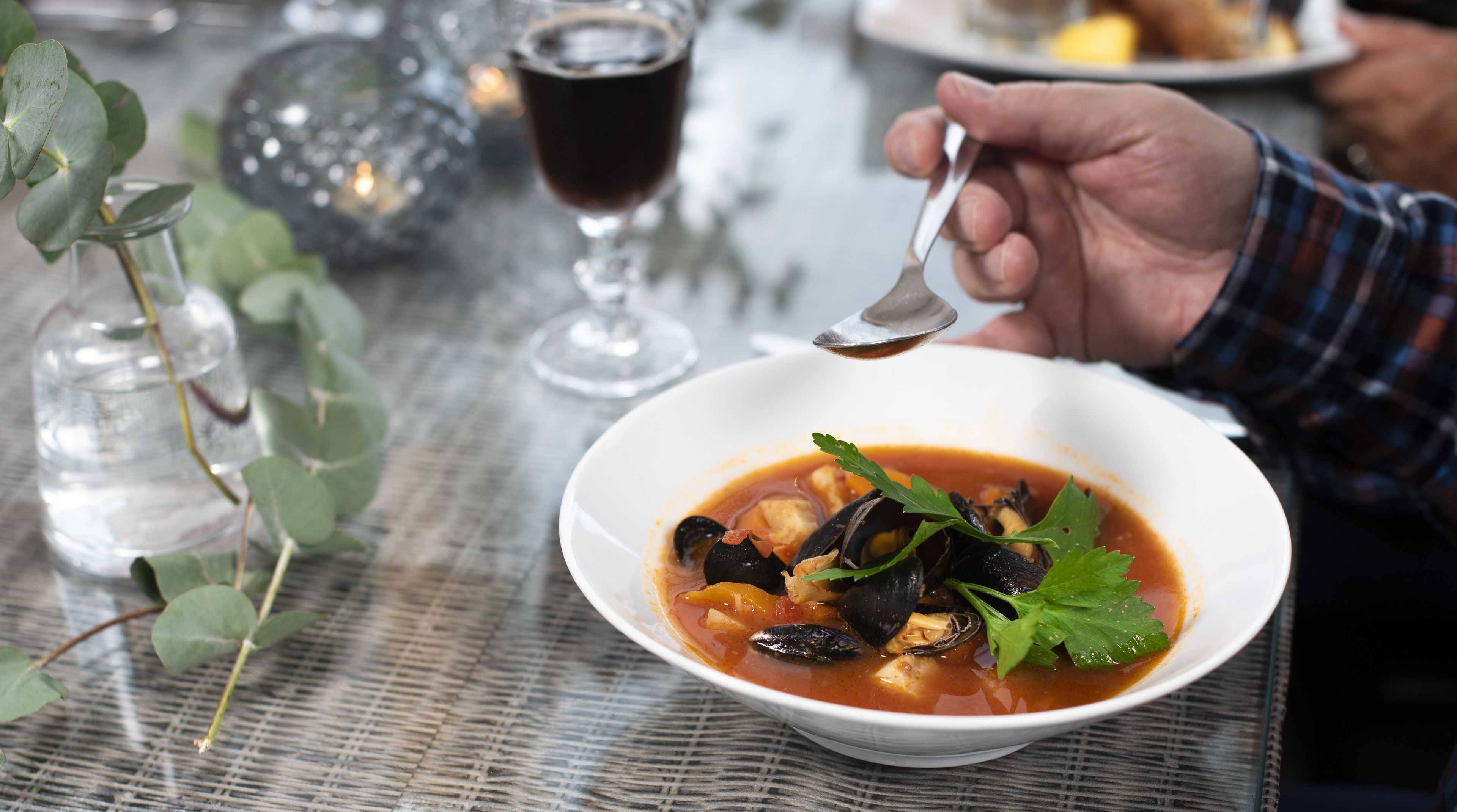 A bowl of seafood soup garnished with parsley, a glass of dark beverage, and a glass of water on a table with a woven placemat.
